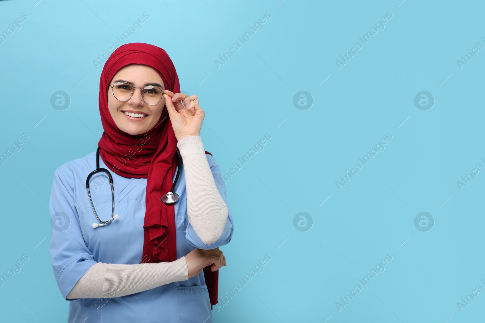 Photo of Muslim woman wearing hijab and medical uniform with stethoscope on light blue background, space for text