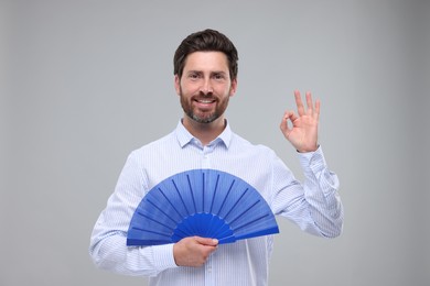 Happy man holding hand fan and showing ok gesture on light grey background