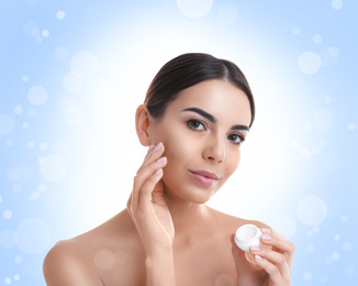 Image of Young woman applying facial cosmetic cream on light blue background with bokeh effect