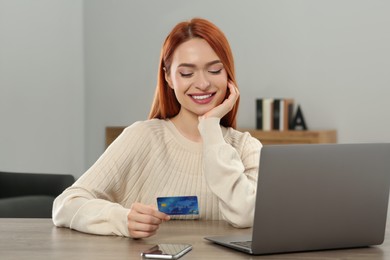 Photo of Happy woman with credit card using laptop for online shopping at wooden table in room