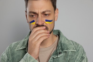Sad man with drawings of Ukrainian flag on face against light grey background, closeup