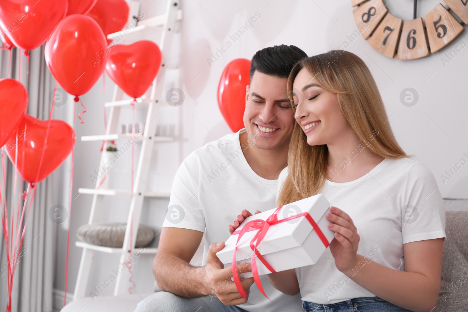 Photo of Man presenting gift to his girlfriend in room decorated with heart shaped balloons. Valentine's day celebration