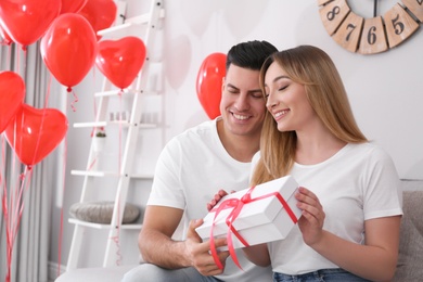 Photo of Man presenting gift to his girlfriend in room decorated with heart shaped balloons. Valentine's day celebration