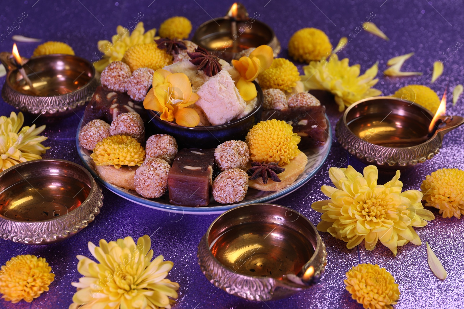 Photo of Diwali celebration. Diya lamps, tasty Indian sweets and chrysanthemum flowers on shiny violet table