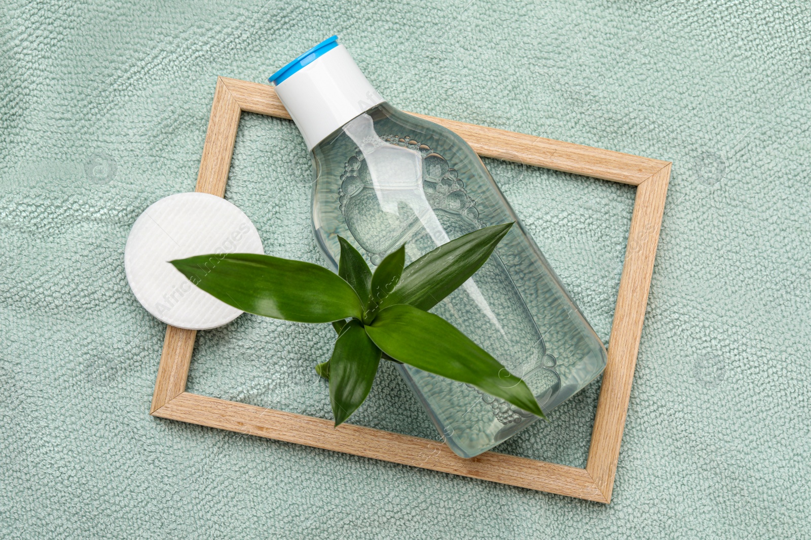 Photo of Bottle of micellar cleansing water, cotton pads and green plant on turquoise towel, flat lay