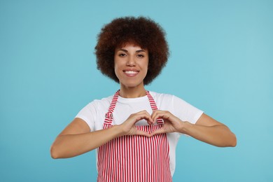 Photo of Happy young woman in apron showing heart gesture on light blue background