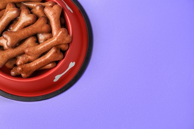 Photo of Red bowl with bone shaped dog cookies on purple background, top view. Space for text