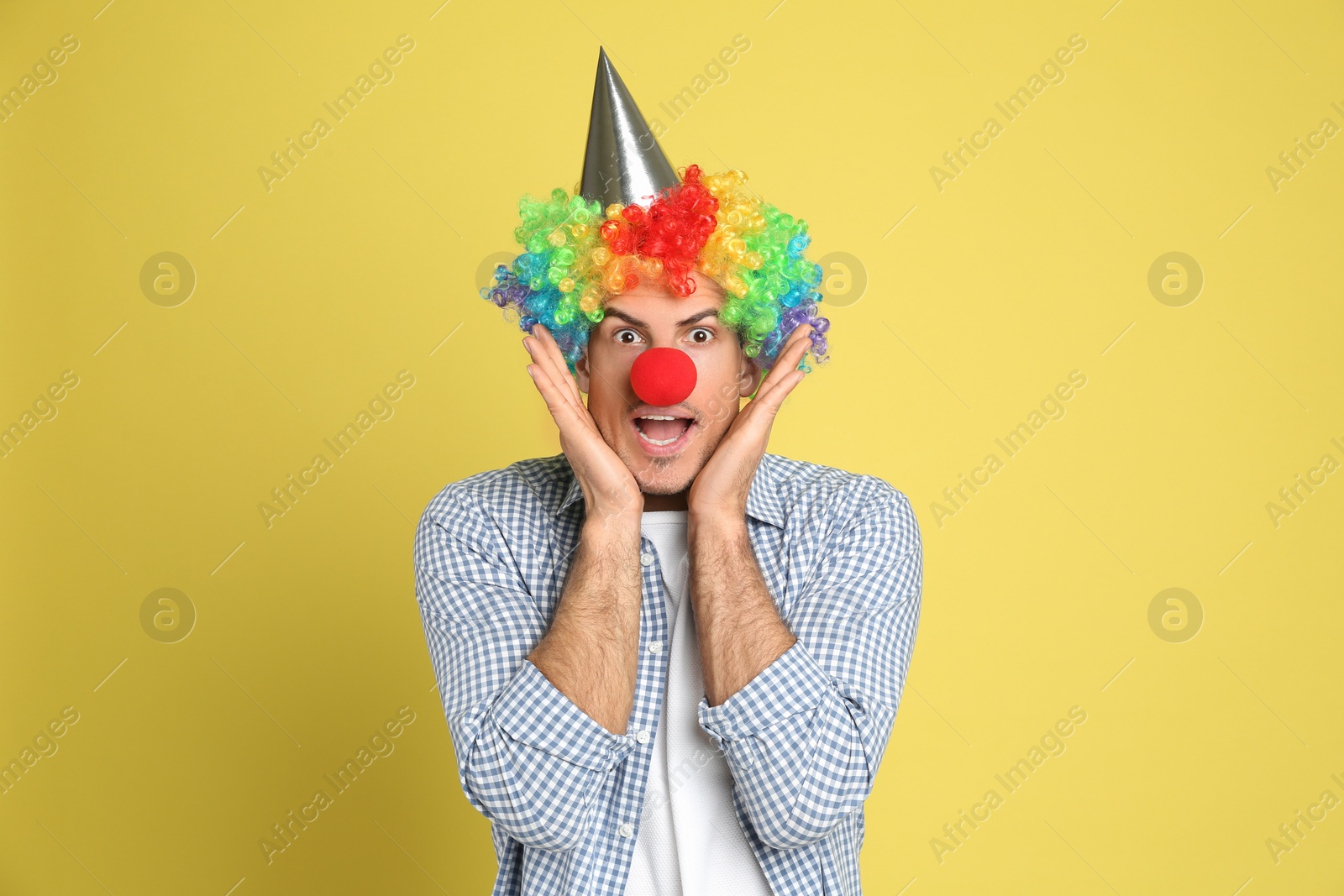 Photo of Funny man with clown nose, party hat and rainbow wig on yellow background. April fool's day