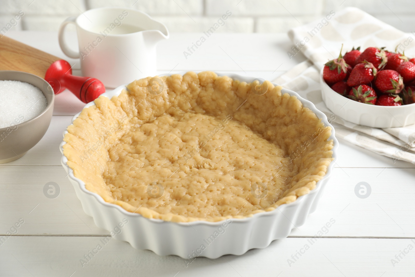 Photo of Making shortcrust pastry. Raw dough in baking dish, strawberries, sugar, rolling pin and milk on white wooden table