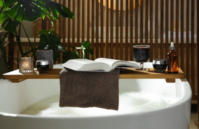 Photo of Wooden bath tray with open book, glass of wine and cosmetic products on tub indoors