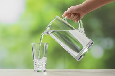 Woman pouring fresh water from jug into glass at white table against blurred green background, closeup