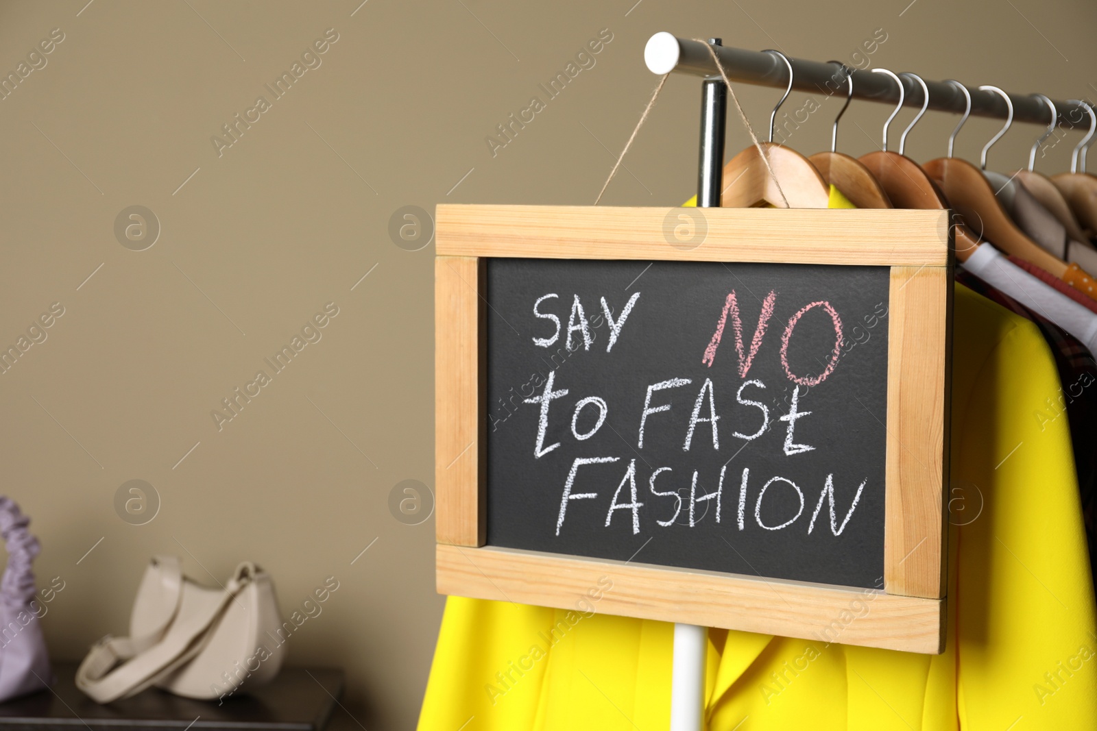 Photo of Small chalkboard with phrase SAY NO TO FAST FASHION and different stylish clothes on rack indoors, space for text