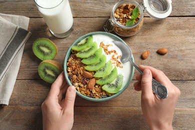 Image of Woman eating tasty granola with yogurt and sliced kiwi for breakfast at wooden table, top view