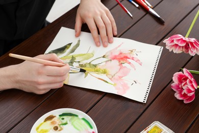 Photo of Woman painting flowers with watercolor at wooden table, closeup. Creative artwork