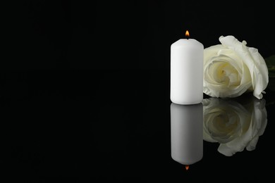 Photo of White rose and burning candle on black mirror surface in darkness, space for text. Funeral symbols
