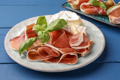 Photo of Slices of tasty cured ham, basil and cheese on blue wooden table, closeup