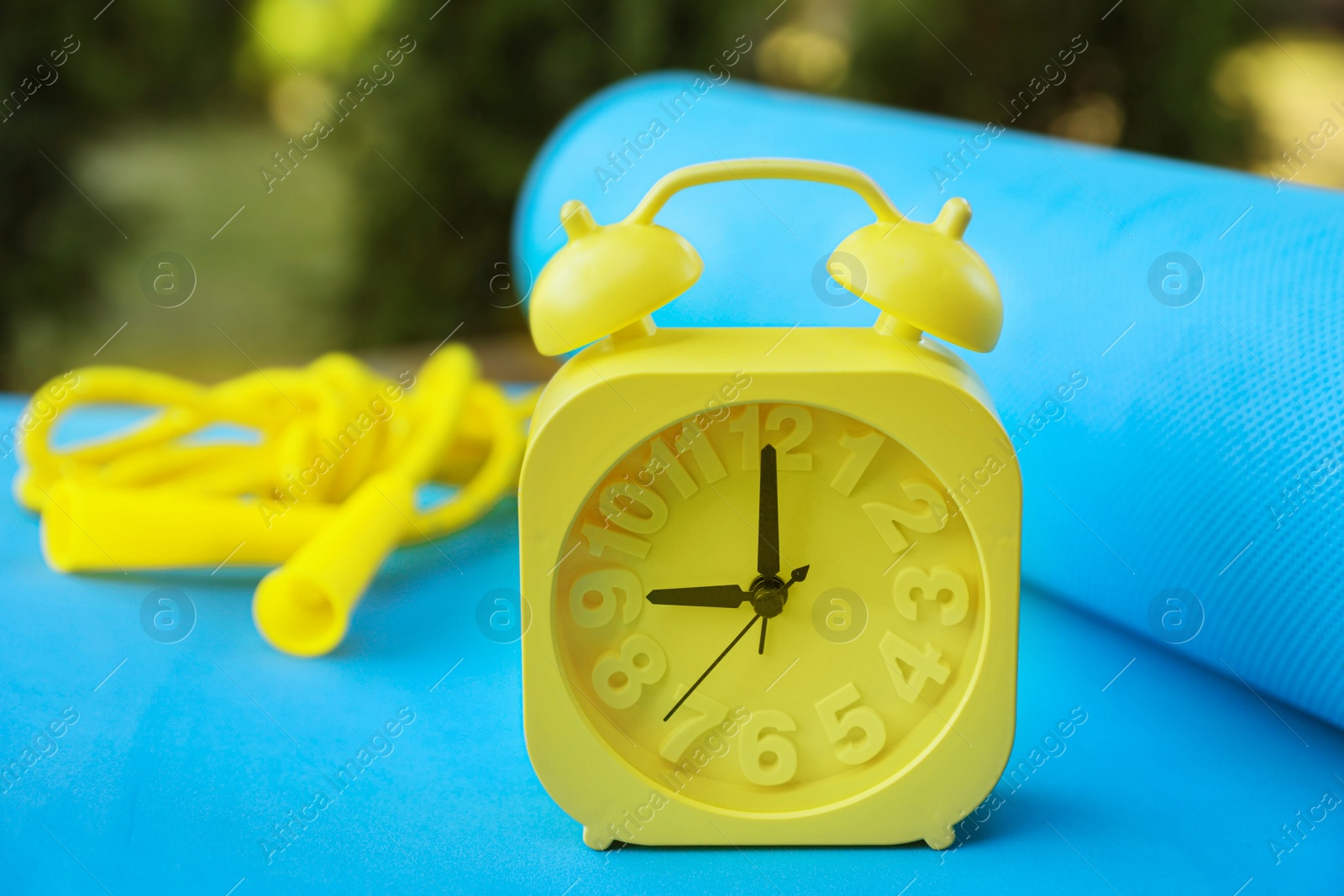 Photo of Alarm clock, fitness mat and skipping rope on wooden table outdoors, closeup. Morning exercise