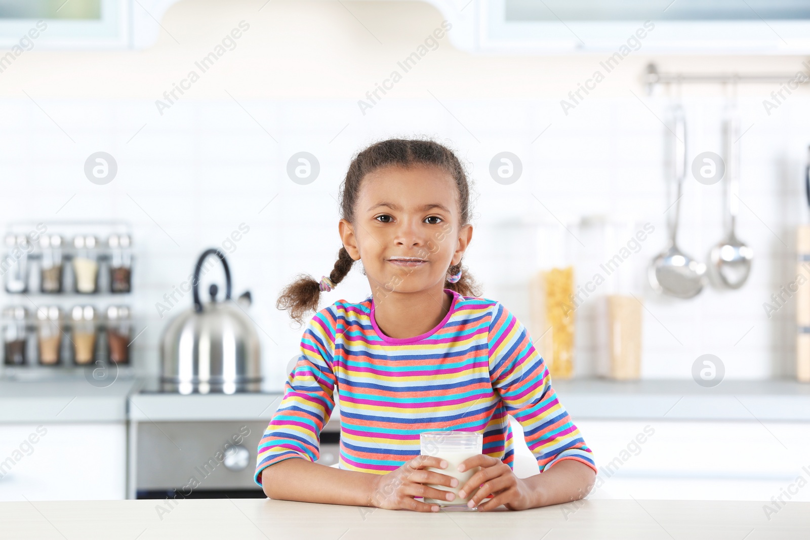 Photo of Adorable African-American girl with glass of milk in kitchen