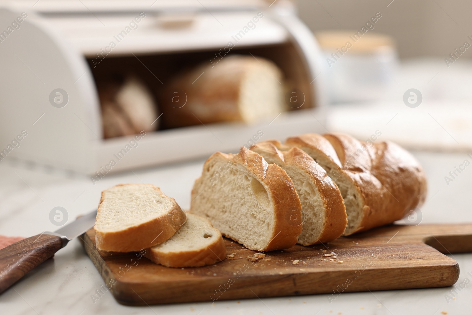 Photo of Wooden bread basket with freshly baked loaves and knife on white marble table in kitchen, closeup