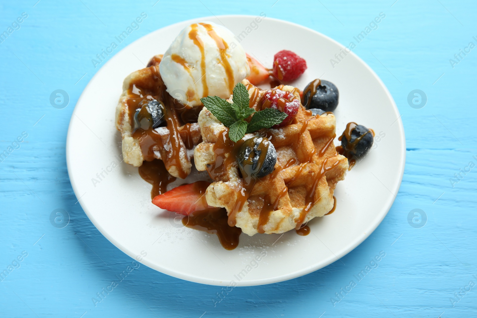 Photo of Delicious Belgian waffles with ice cream, berries and caramel sauce on light blue wooden table, closeup