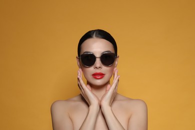 Attractive woman in fashionable sunglasses touching face against orange background