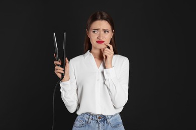 Photo of Upset young woman with flattening iron on black background. Hair damage