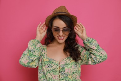 Beautiful young woman with straw hat and stylish sunglasses on pink background