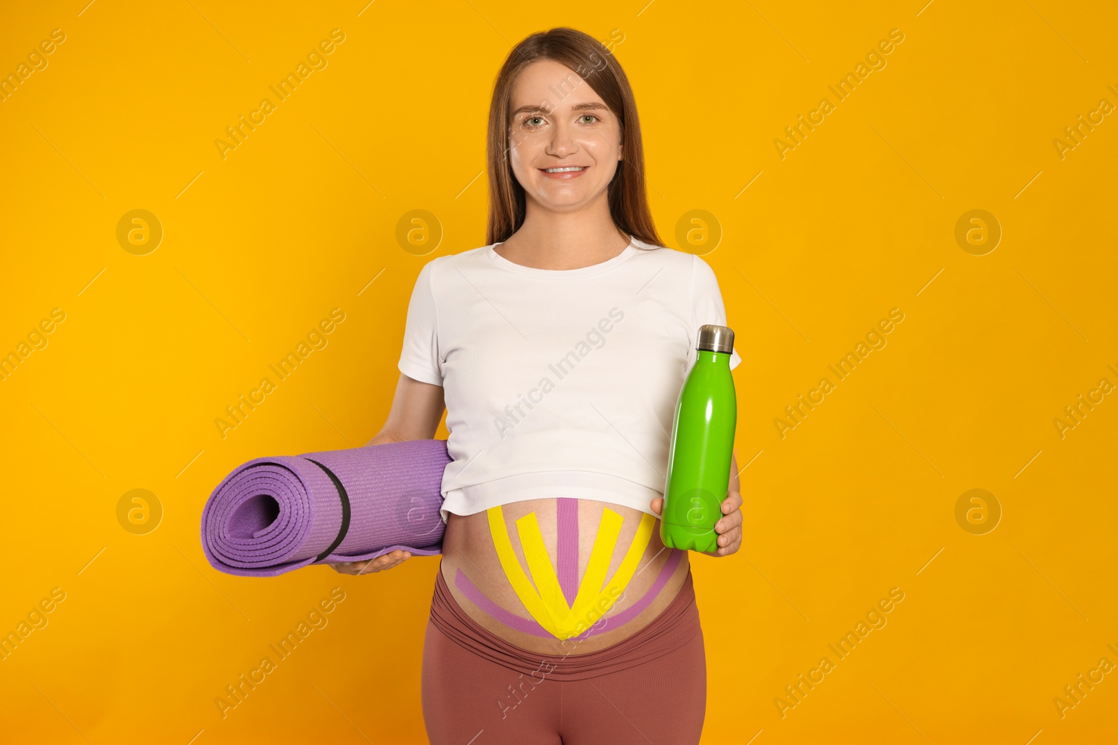 Photo of Beautiful pregnant woman with kinesio tapes holding water bottle and mat on orange background