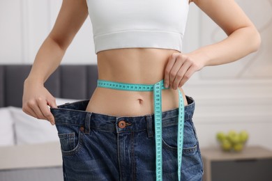 Photo of Slim woman wearing big jeans and measuring waist with tape in room, closeup. Weight loss