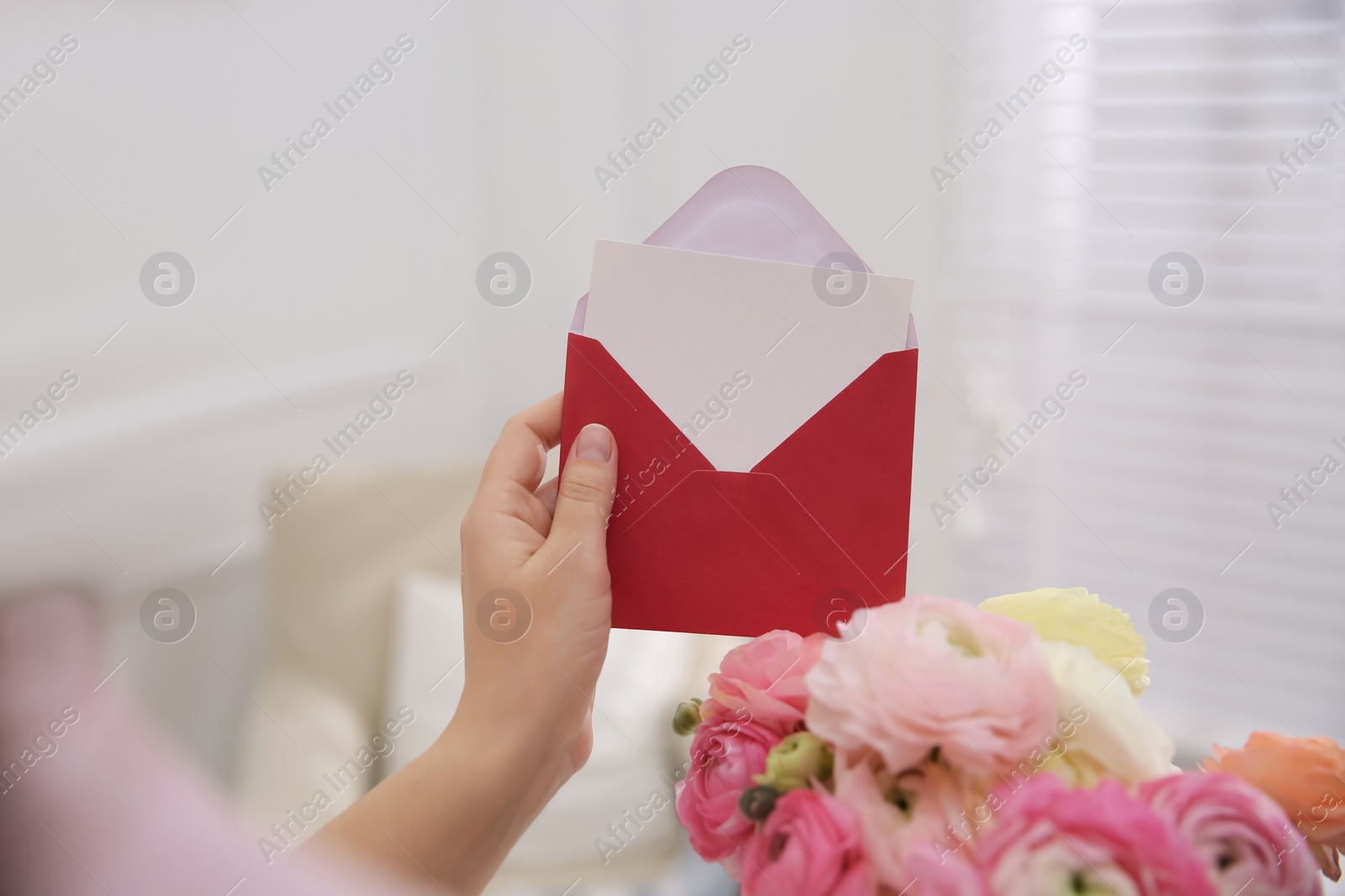 Photo of Woman holding envelope with blank greeting card and bouquet of ranunculus at home, closeup