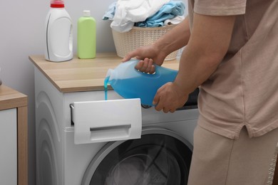 Photo of Man pouring fabric softener from bottle into washing machine indoors, closeup