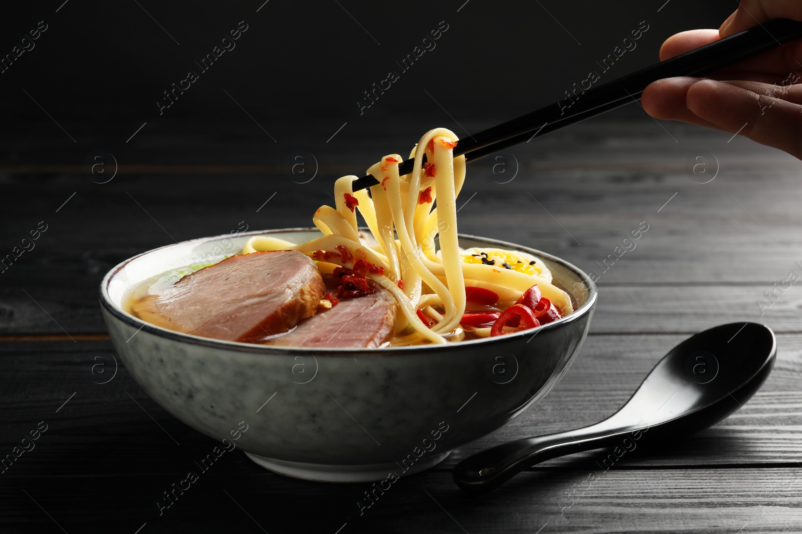 Photo of Woman eating delicious ramen with chopsticks at black wooden table, closeup. Noodle soup