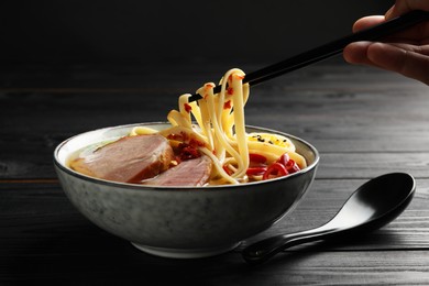 Woman eating delicious ramen with chopsticks at black wooden table, closeup. Noodle soup