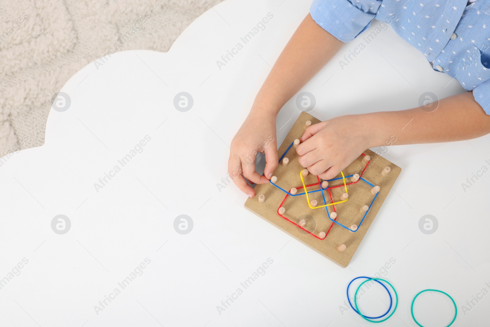Photo of Motor skills development. Girl playing with geoboard and rubber bands at white table, top view. Space for text
