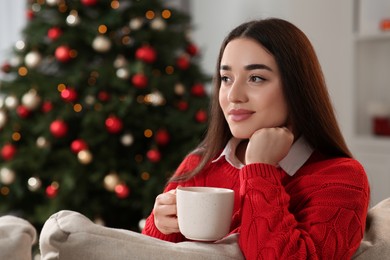 Woman holding cup of hot drink on sofa near Christmas tree indoors. Space for text