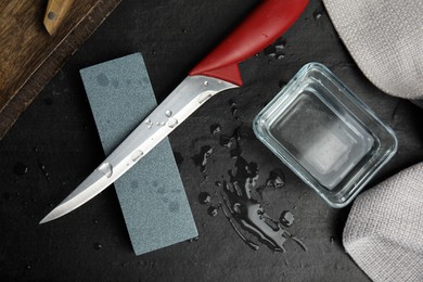 Sharpening stone, knife and water on black table, flat lay