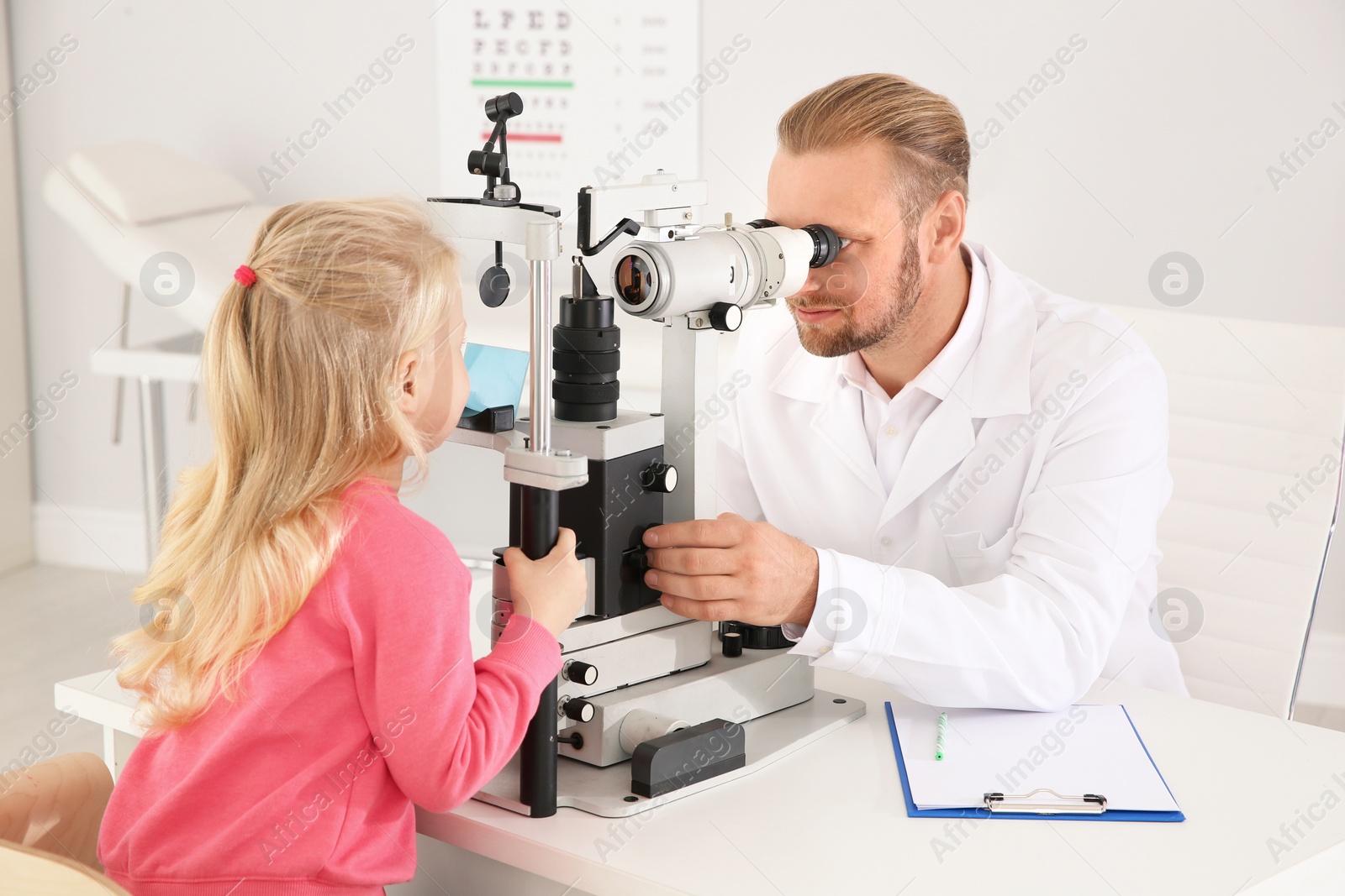 Photo of Children's doctor examining little girl with ophthalmic equipment in clinic