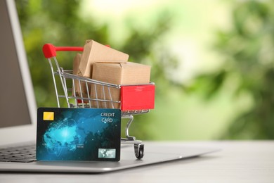 Online payment concept. Small shopping cart with bank card, boxes and laptop on white table, space for text