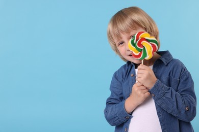 Little boy covering face with colorful lollipop swirl on light blue background, space for text
