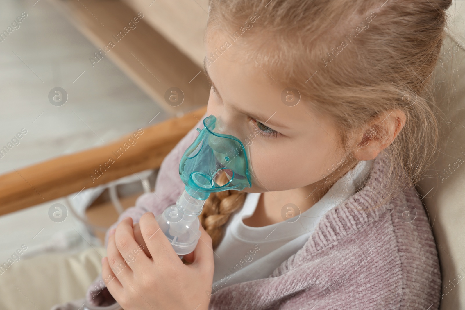 Photo of Little girl using nebulizer for inhalation in armchair at home