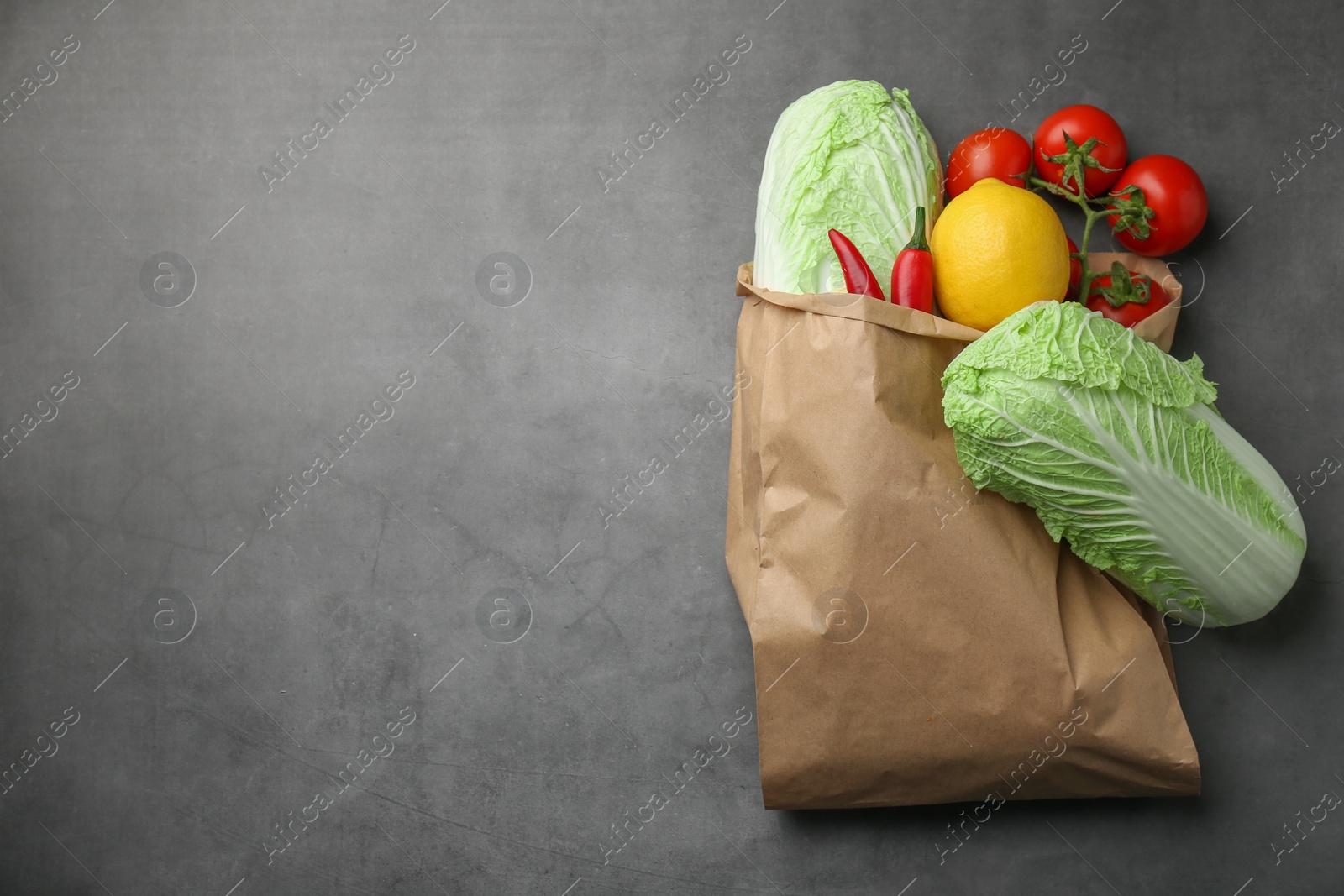 Photo of Paper bag with fresh Chinese cabbages, lemon, tomatoes and chili pepper on grey textured table, top view. Space for text