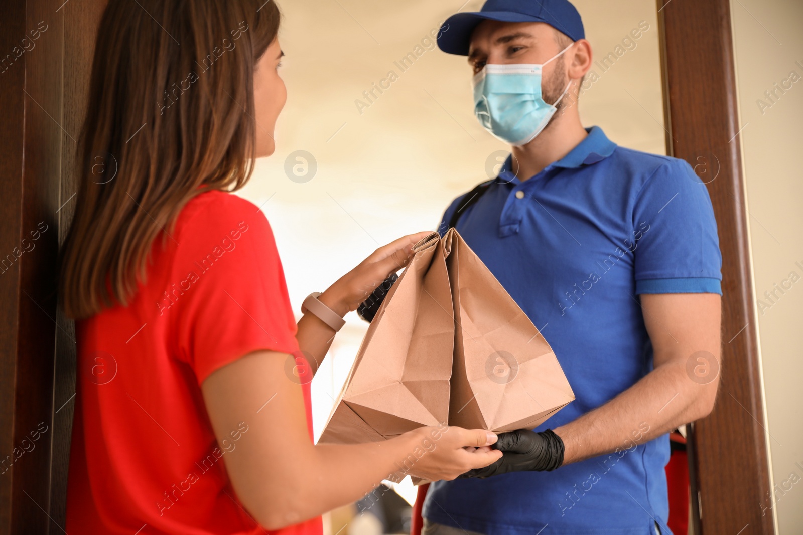 Photo of Courier in protective mask and gloves giving order to woman at entrance. Restaurant delivery service during coronavirus quarantine