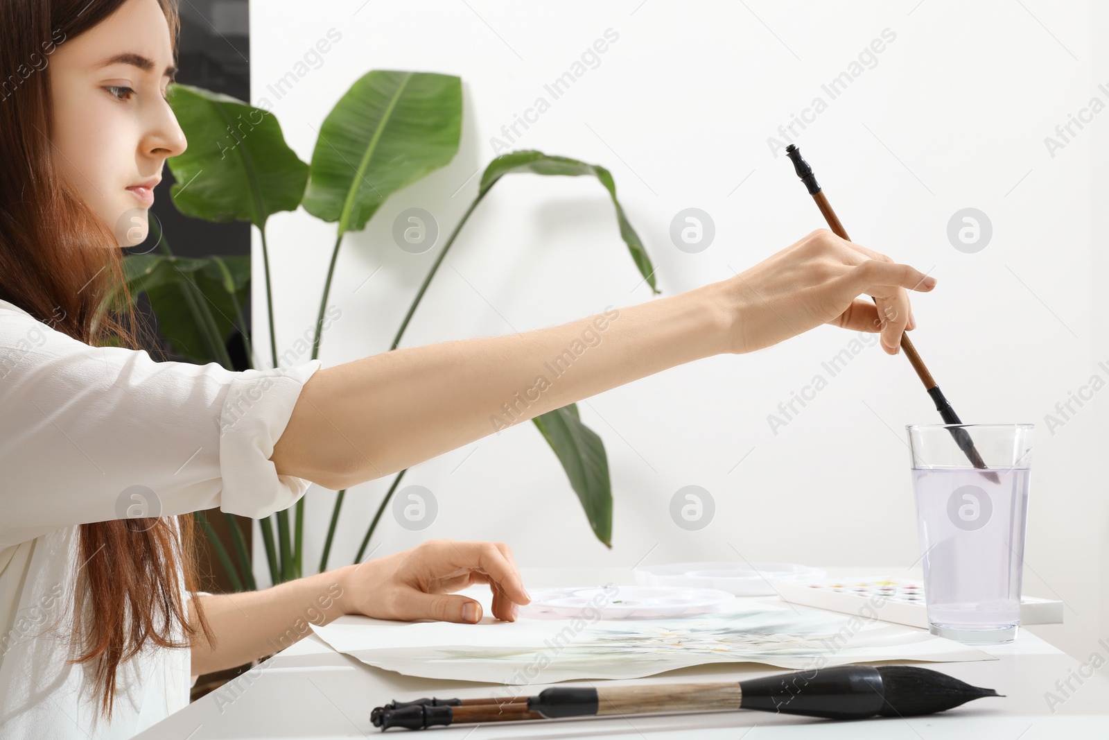 Photo of Woman wetting brush at white table indoors. Watercolor artwork