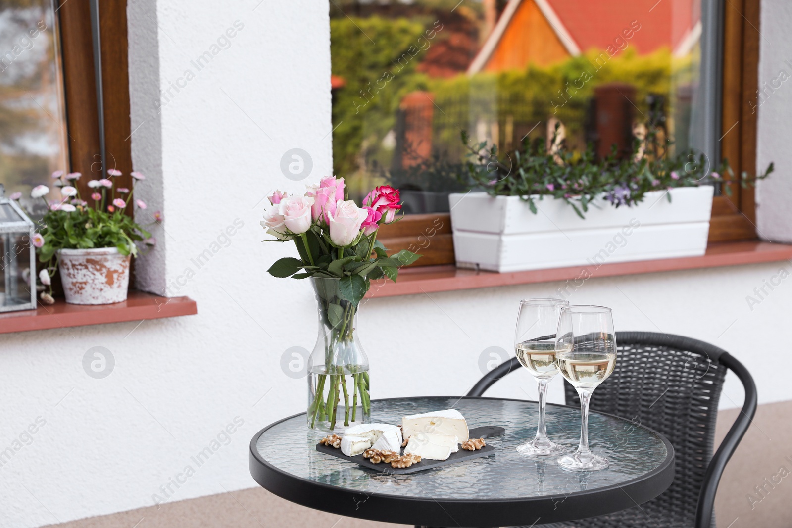 Photo of Vase with roses, glasses of wine and food on glass table near house on outdoor terrace