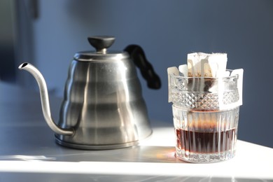Photo of Glass with drip coffee bag and kettle on white table, closeup