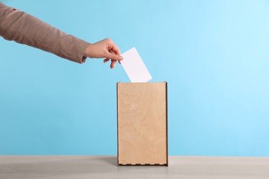 Photo of Woman putting her vote into ballot box on wooden table against light blue background, closeup