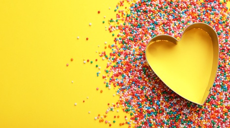 Photo of Heart shaped cookie cutter and sprinkles on yellow background, flat lay with space for text. Confectionery decor