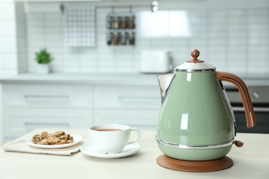 Modern electric kettle, cup of tea and cookies on wooden table in kitchen