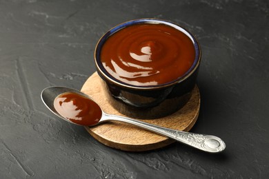 Photo of Tasty barbeque sauce in bowl and spoon on dark textured table, closeup