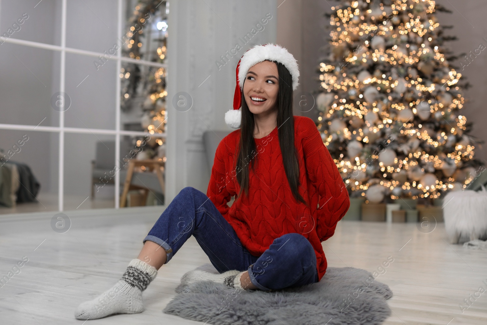 Photo of Happy young woman wearing Santa hat in room with Christmas tree